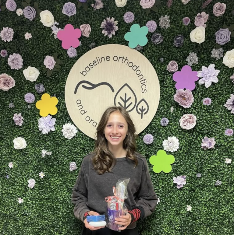Smiling young girl holding dental items while standing in front of a decorative flower wall with the Baseline Orthodontics and Oral Surgery logo.