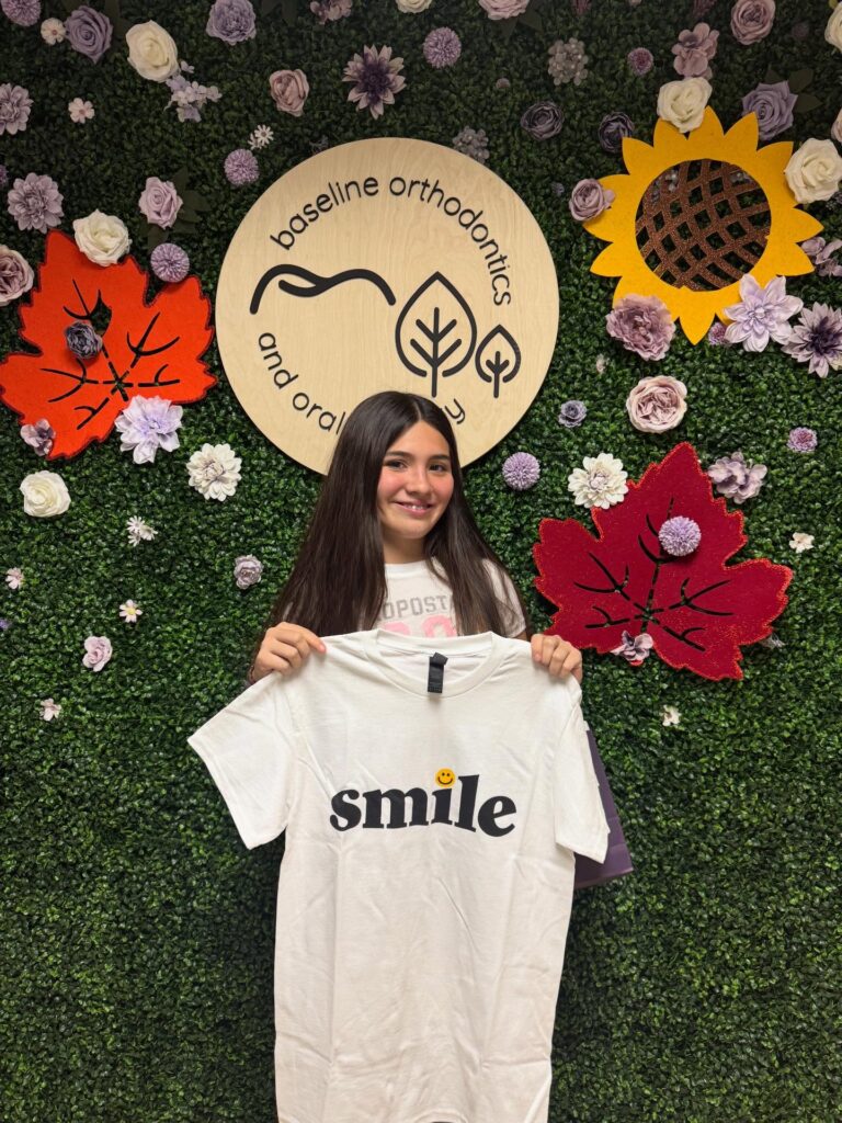 Smiling young patient holding a white ‘smile’ T-shirt while standing in front of a decorative flower wall with the Baseline Orthodontics and Oral Surgery logo.