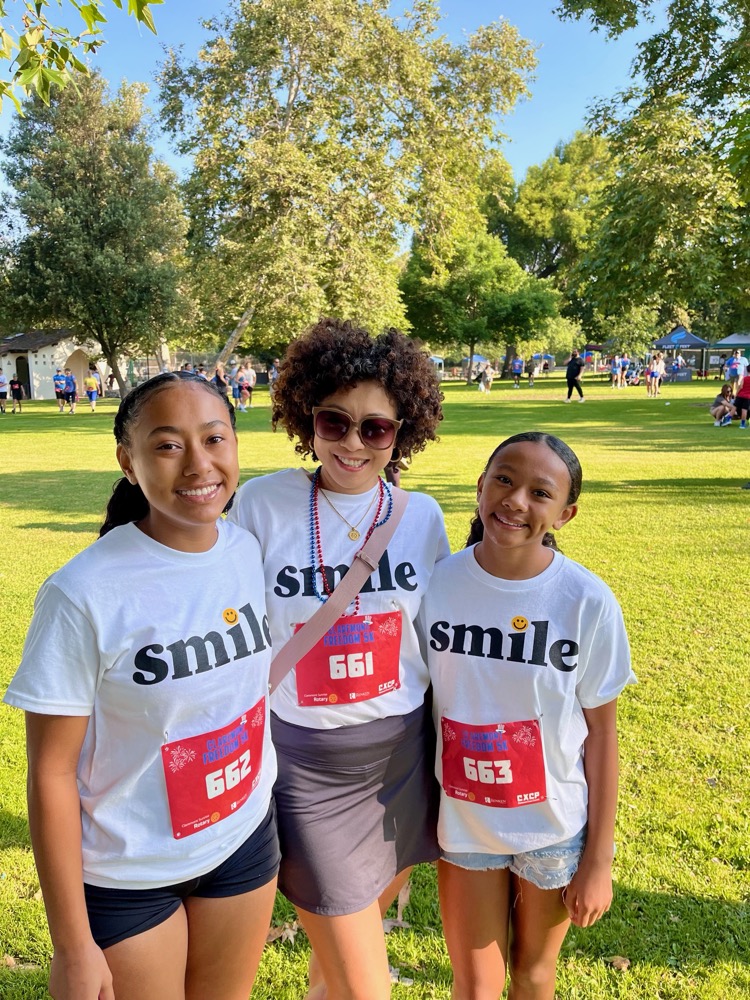 Three smiling people wearing matching white ‘smile’ shirts and race bibs posing together at an outdoor community event in a park.