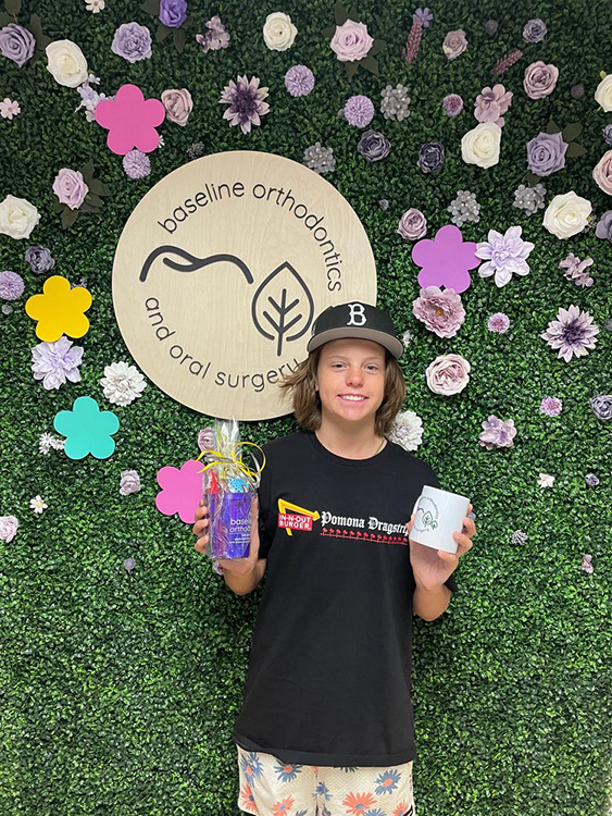 Smiling young patient holding gifts while standing in front of a decorative flower wall with the Baseline Orthodontics and Oral Surgery logo.