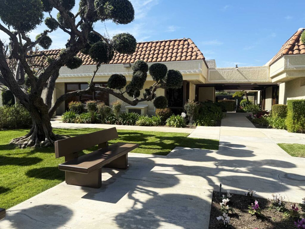 Landscaped courtyard outside a Spanish-style building with trimmed topiary trees, green lawn, walkway, and a bench under a sunny blue sky.
