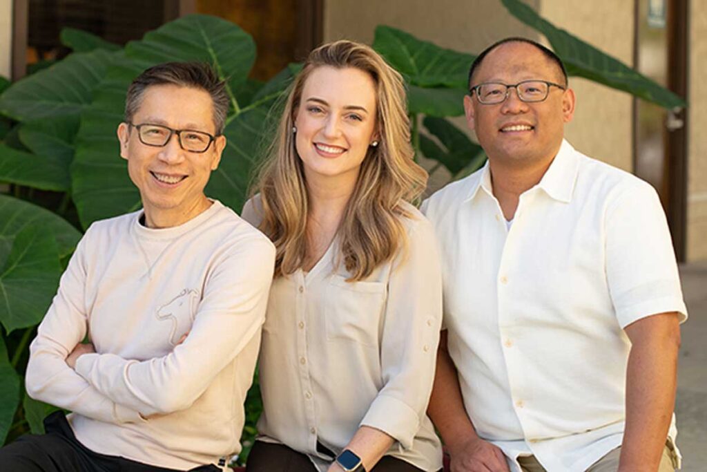 The expert orthodontic team at Baseline Orthodontics smiling together outdoors. Two male orthodontists and one female orthodontist sit side-by-side in front of green foliage, showcasing their friendly and professional dental care staff.