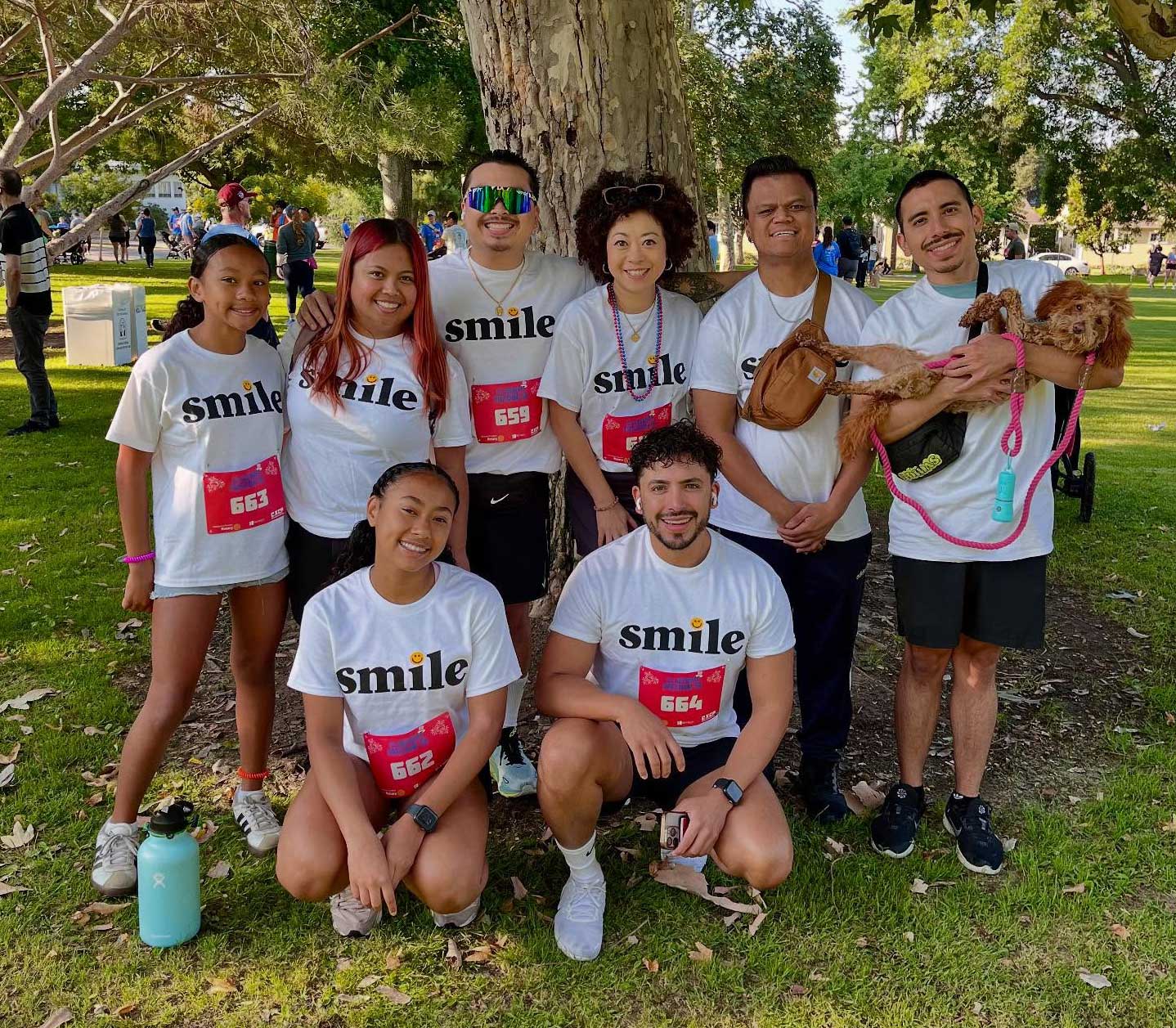 Group of eight people in white 'smile' shirts posing together in a park, with a small brown dog being held by one member on the right.